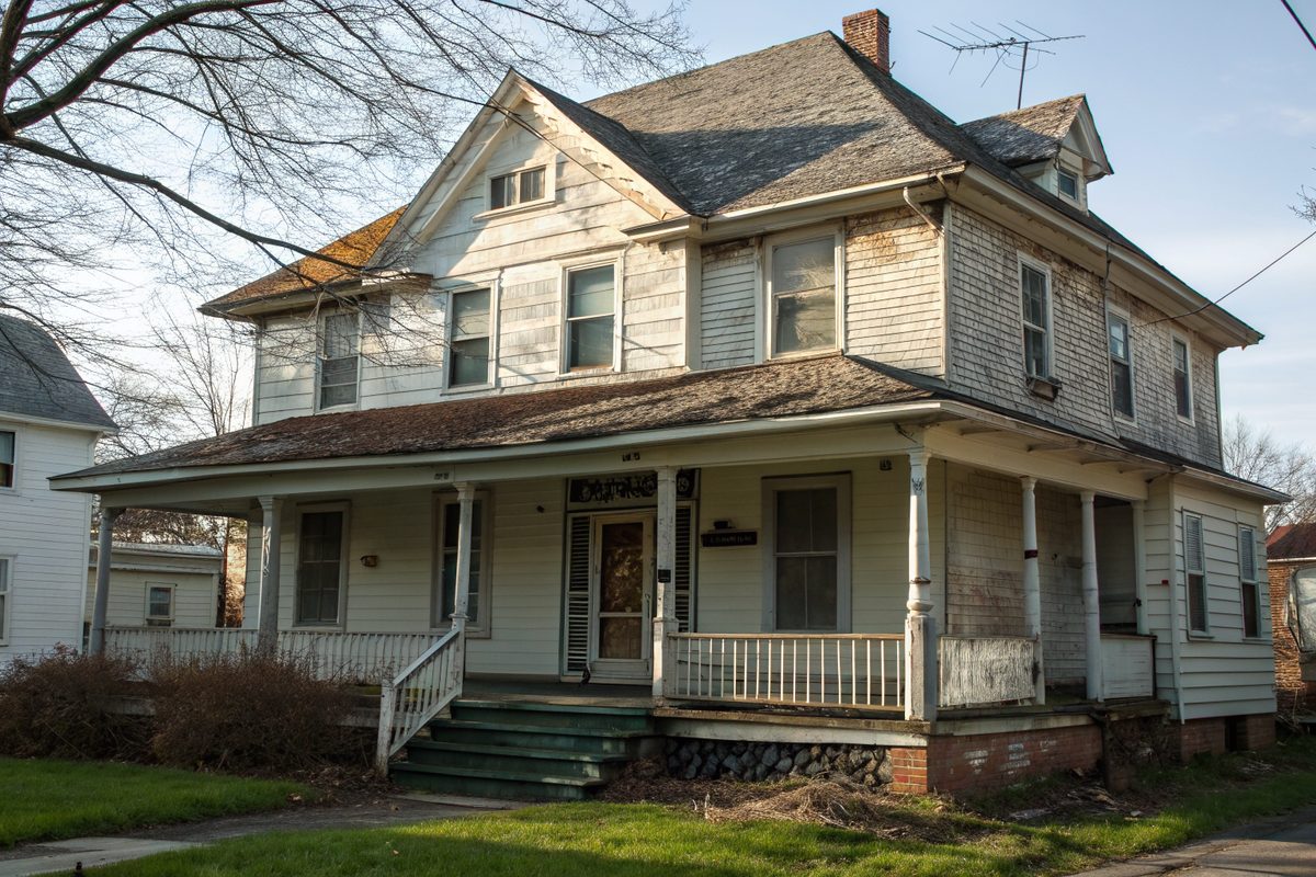 An older frame twin house with a porch and a worn, mismatched shingle roof