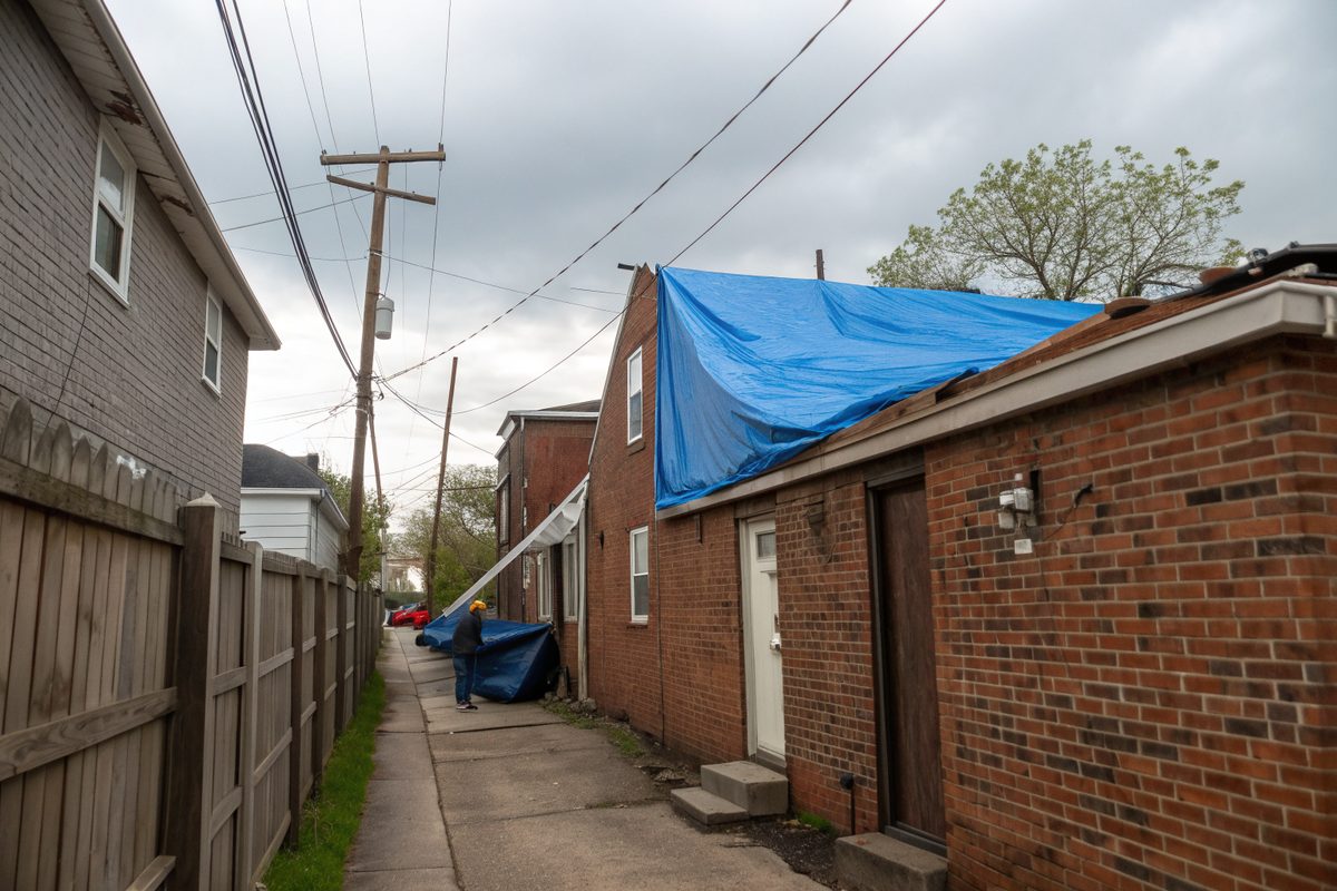 A blue tarp held by boards over the flat rear roof of a brick row home